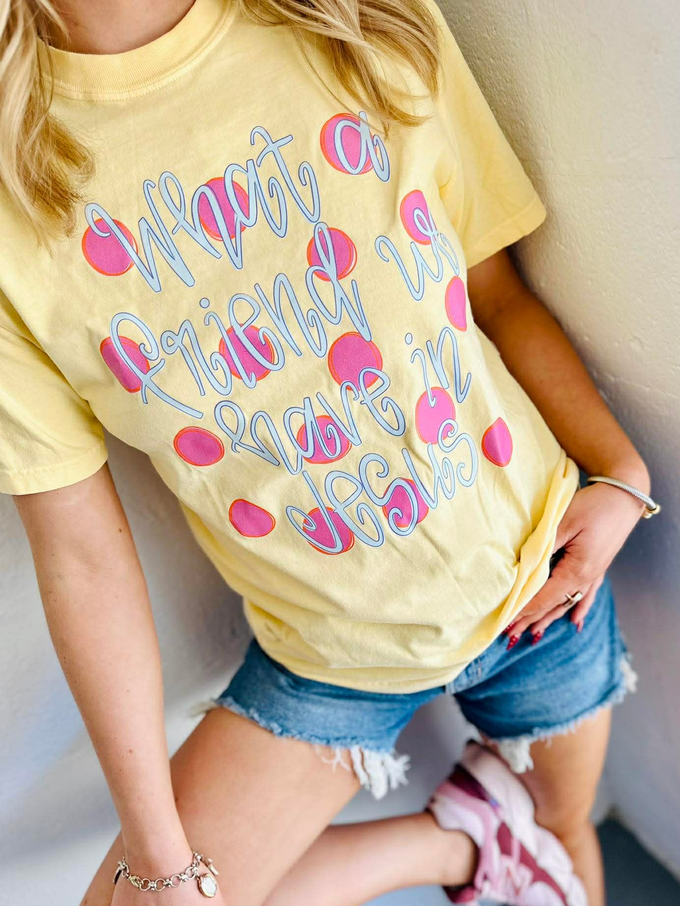 Person wearing a yellow t-shirt with text "What a friend we have in Jesus" polka dots and pink hearts, sitting against a light-colored wall.