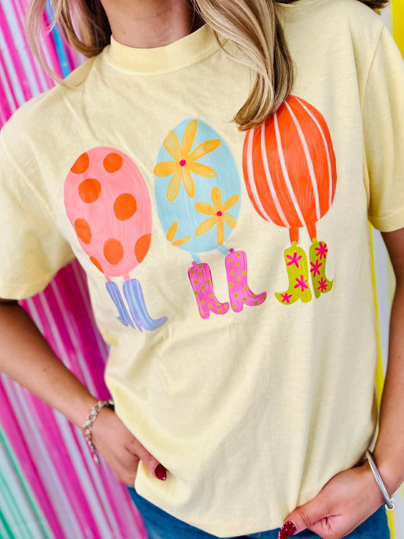 Person wearing a yellow t-shirt with colorful egg and boots balloon designs on a multicolored striped background