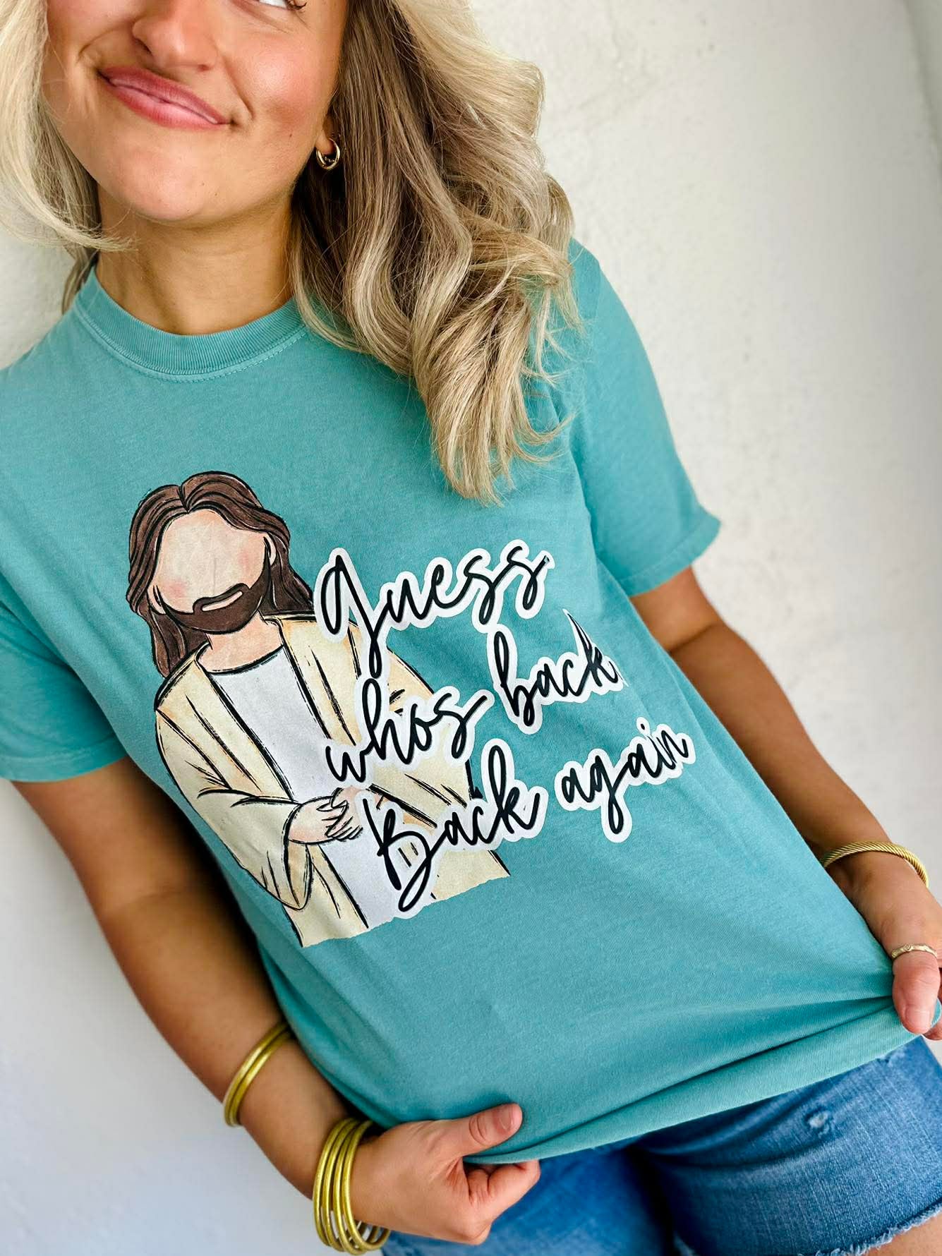 Person wearing a teal t-shirt with a Jesus "guess who is back again" graphic design and text.