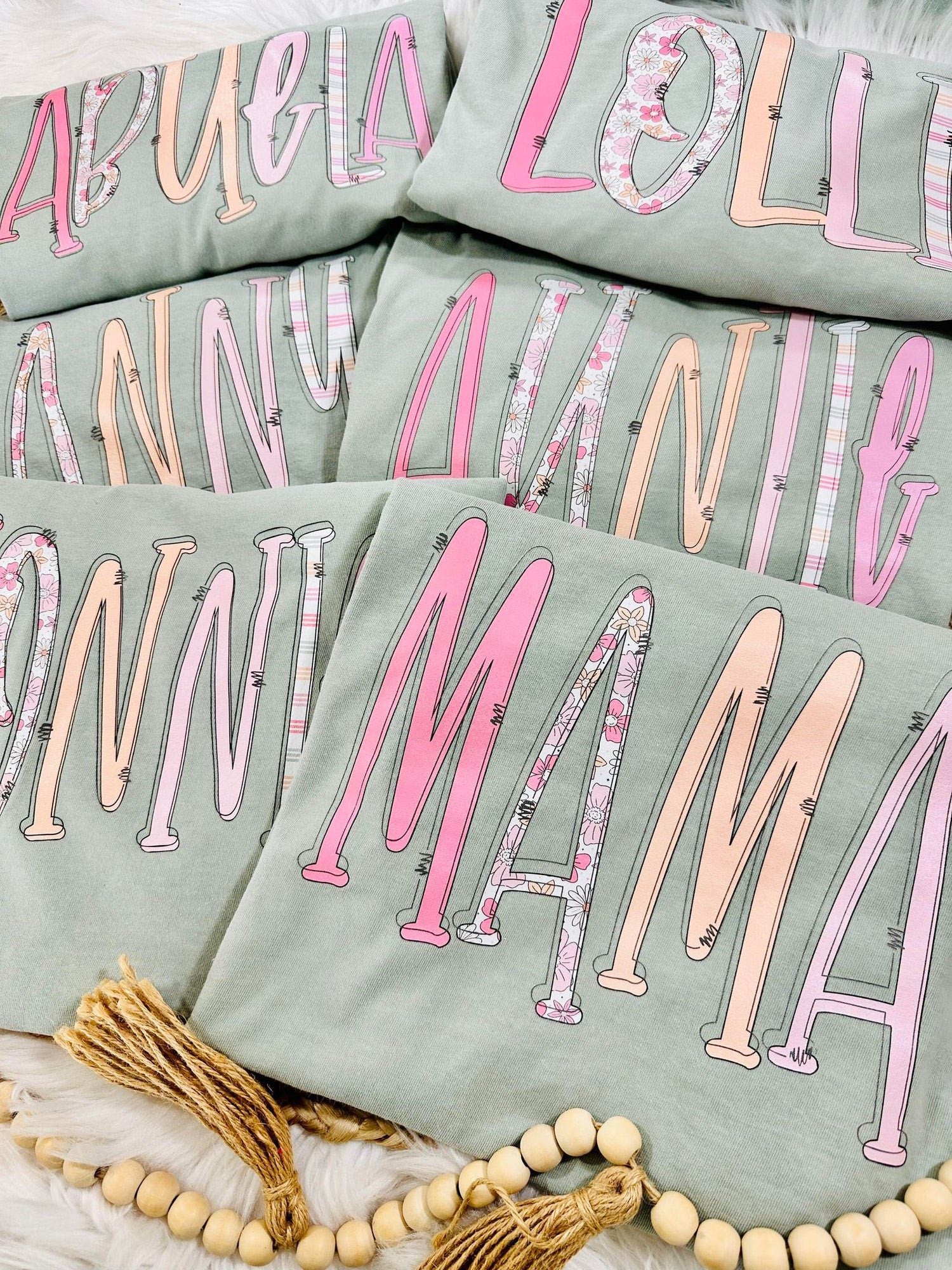 Stack of Bay Comfort Color tees with pink and white MAMA AUNTIE NONNI ABUELA NANNY LOLLI text on a light surface.