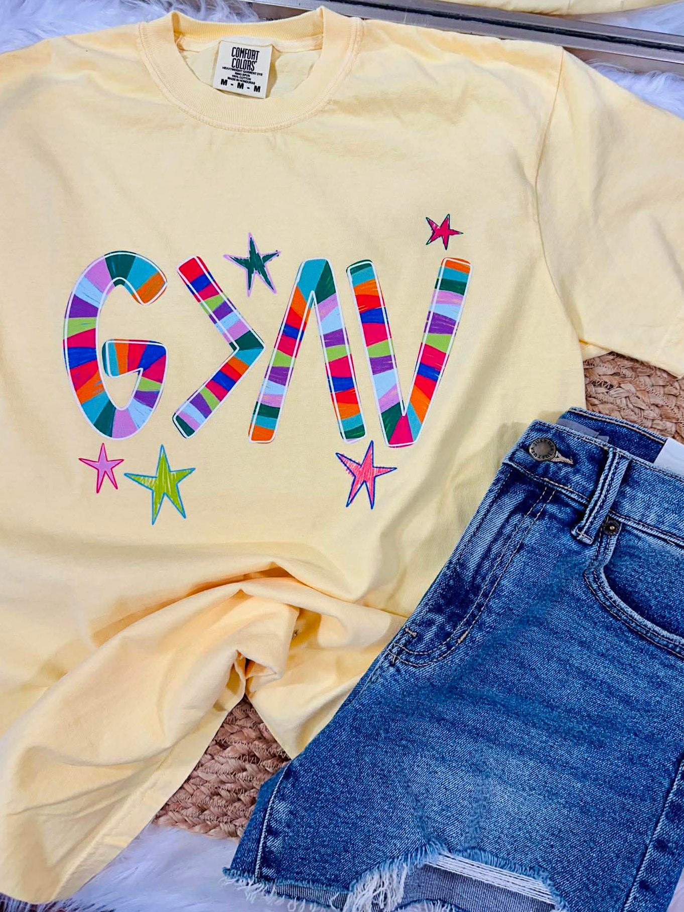 Yellow t-shirt with colorful 'G>^V' text and denim shorts on a textured surface