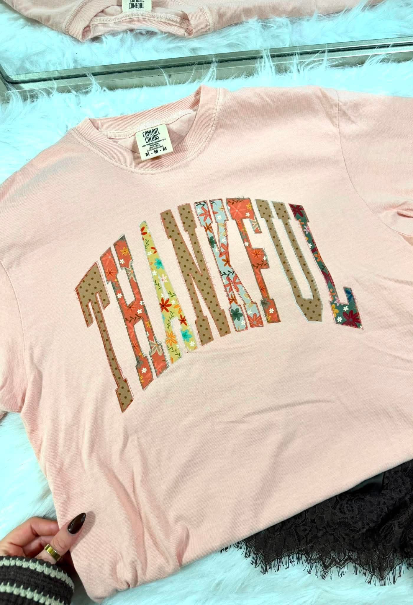 A light pink long-sleeve t-shirt with the word 'THANKFUL' printed across the chest in a retro arched font filled with multicolor floral and patterned fabrics. A hand is visible holding the bottom left corner of the shirt, which is laid out on a white faux fur surface.