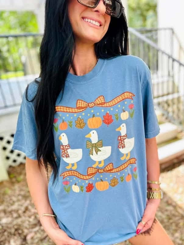 A woman wearing a light blue graphic t-shirt with a fall-themed design of white ducks, pumpkins, and autumn leaves. She is posing outdoors in front of a porch staircase.