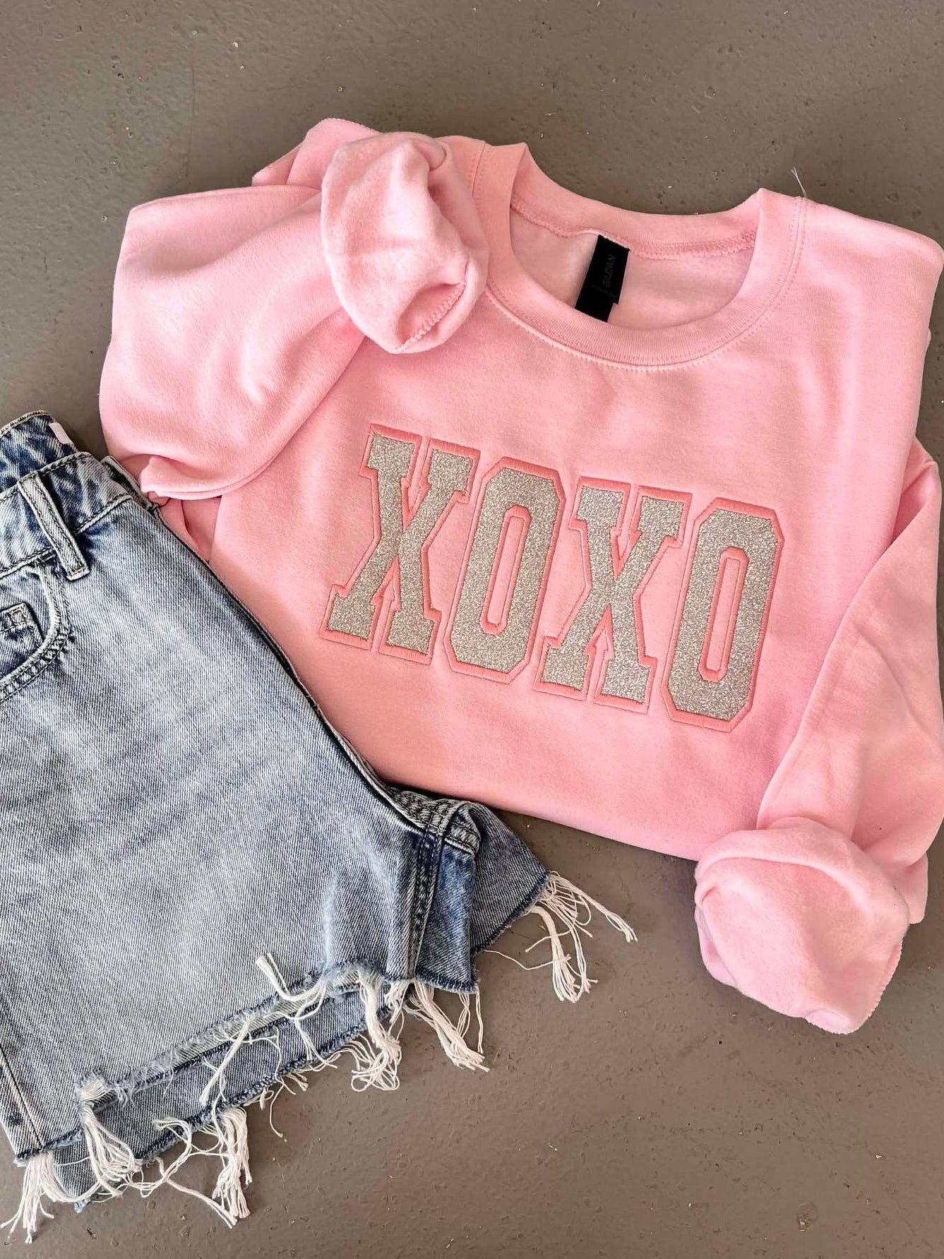 Pink sweatshirt with 'XOXO' Glitter Applique embroidery and denim shorts on a gray background