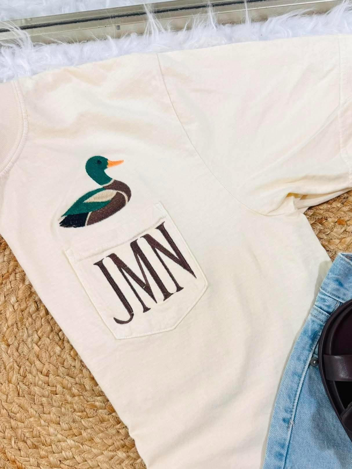 An off-white short-sleeve t-shirt with a front pocket featuring a duck graphic and the monogram 'JMN', styled in a flat lay with light-wash denim shorts and dark brown slide sandals on a woven rug.