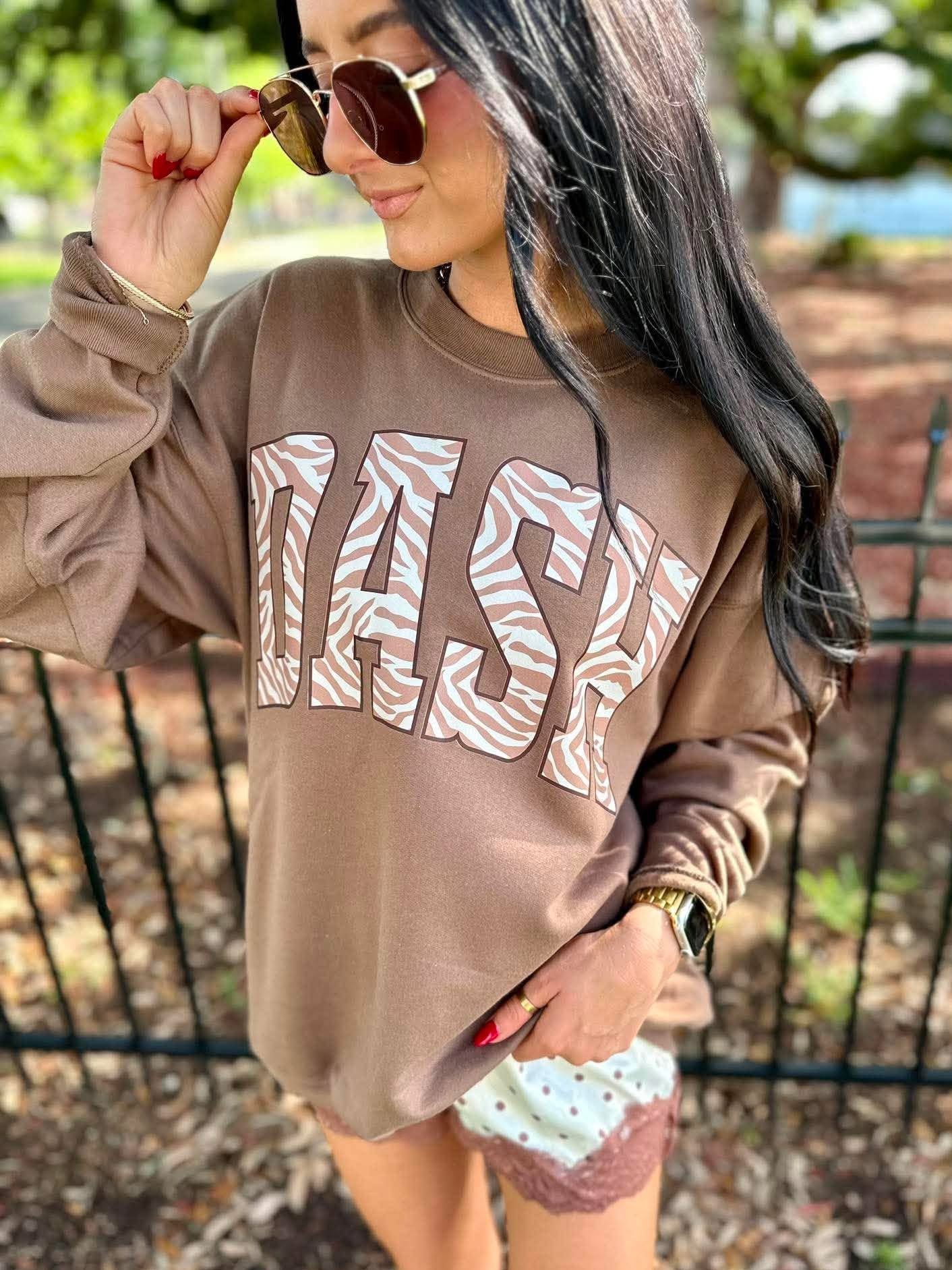 A woman wearing a brown sweatshirt with 'DASH' printed in large white and tan zebra-patterned letters, paired with patterned shorts and sunglasses, standing outdoors in front of a fence.