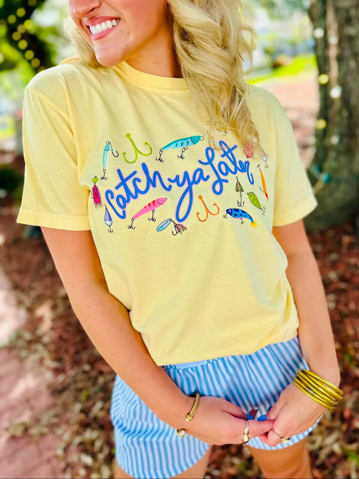 A woman wearing a pale yellow graphic t-shirt with fishing lures and the text 'catch ya later' in blue script, paired with blue and white striped shorts, standing outdoors in front of a tree with string lights.
