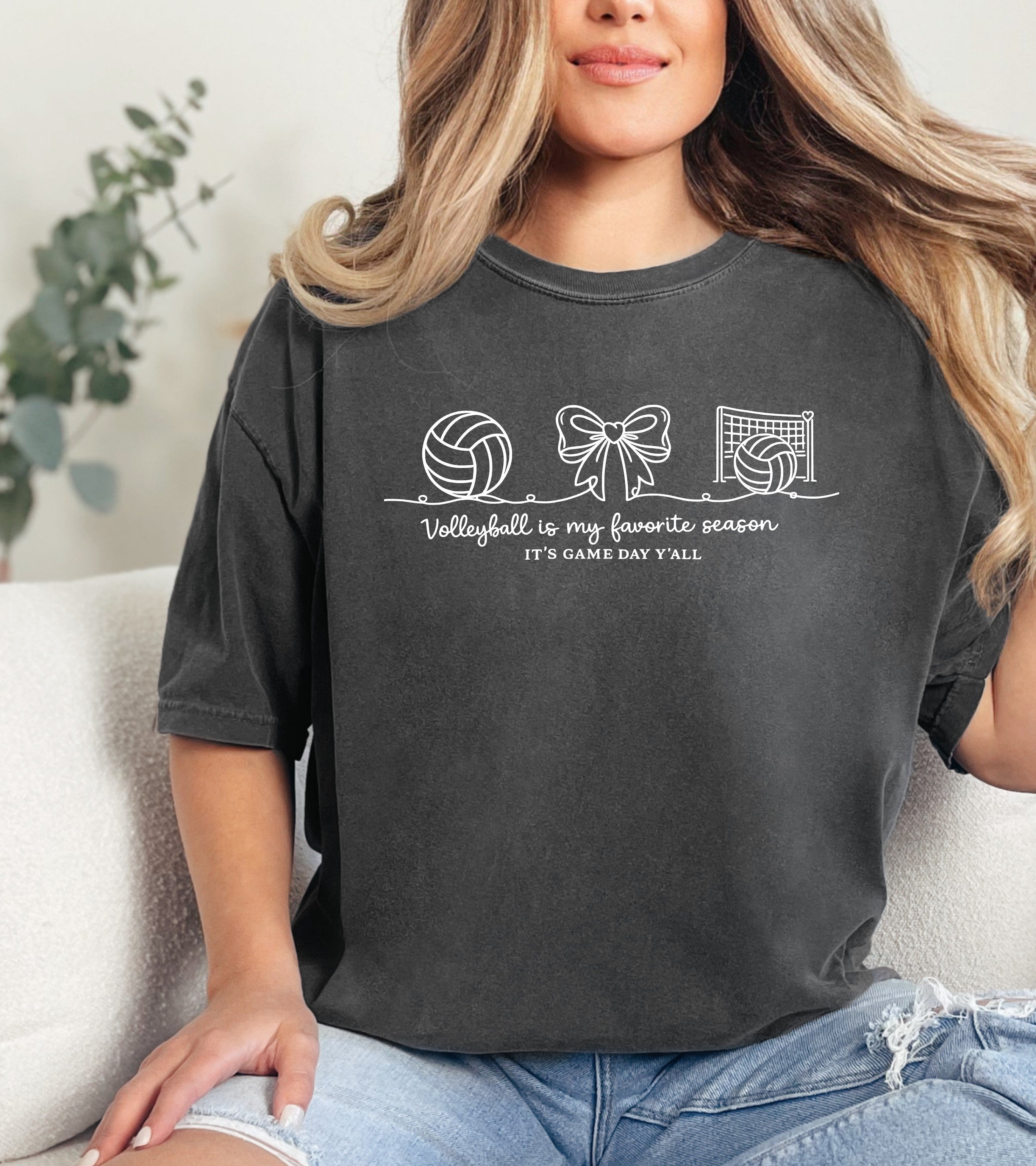 Person wearing a dark gray t-shirt with sports-themed graphics and Person wearing a dark gray t-shirt with a "Volleyball is my favorite season" graphic design and text, sitting on a couch. text.