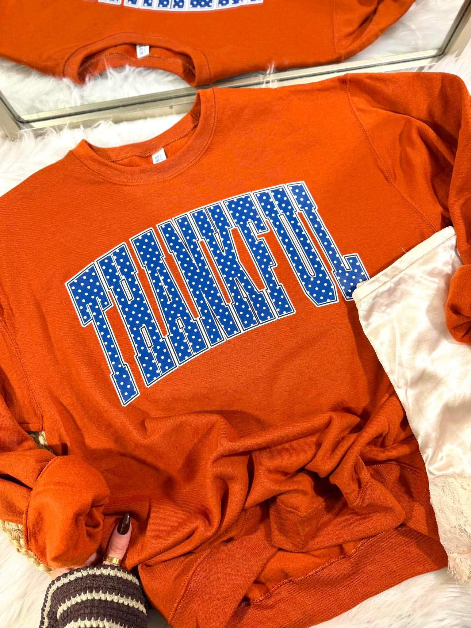 An orange crewneck sweatshirt with 'THANKFUL' written across the chest in blue block letters filled with white stars, styled on a white faux fur surface.