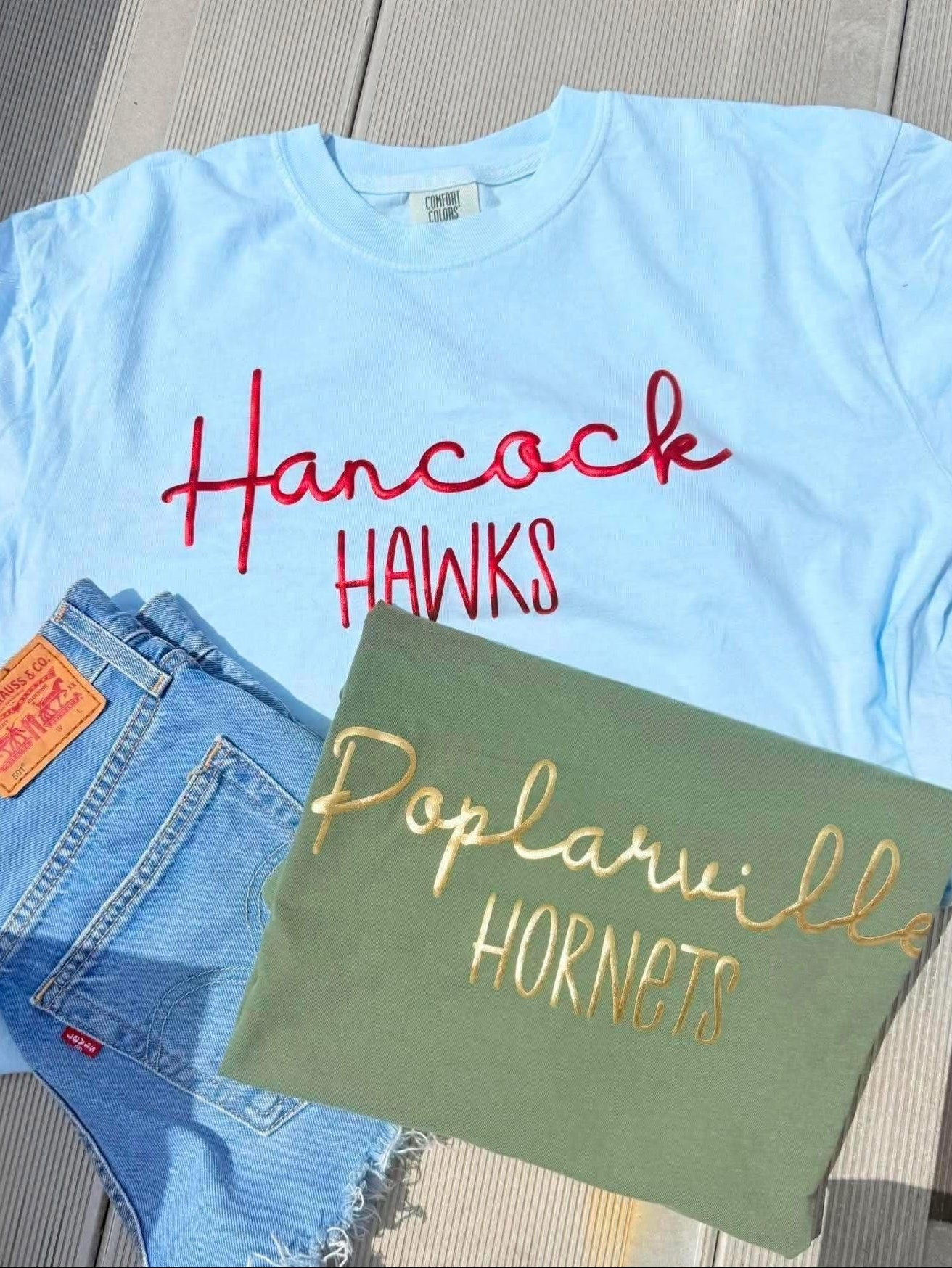 Light blue t-shirt with 'Hancock Hawks' text, Levi's jeans, and green tote bag with 'Poplarville Hornets' text on a wooden surface.