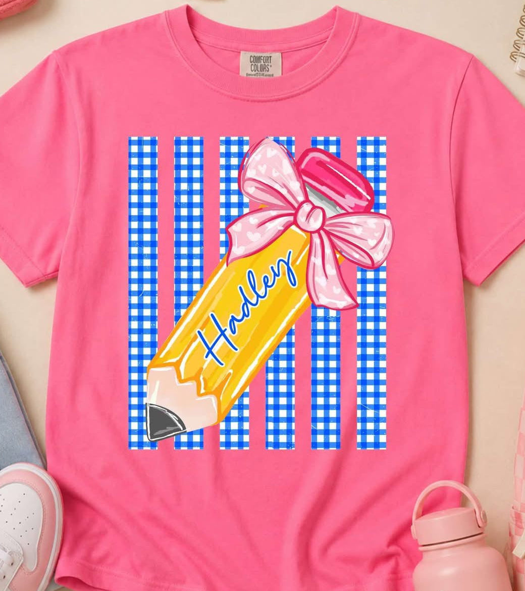Pink t-shirt with a pencil graphic and 'Haley' text, surrounded by stationery items on a light background.