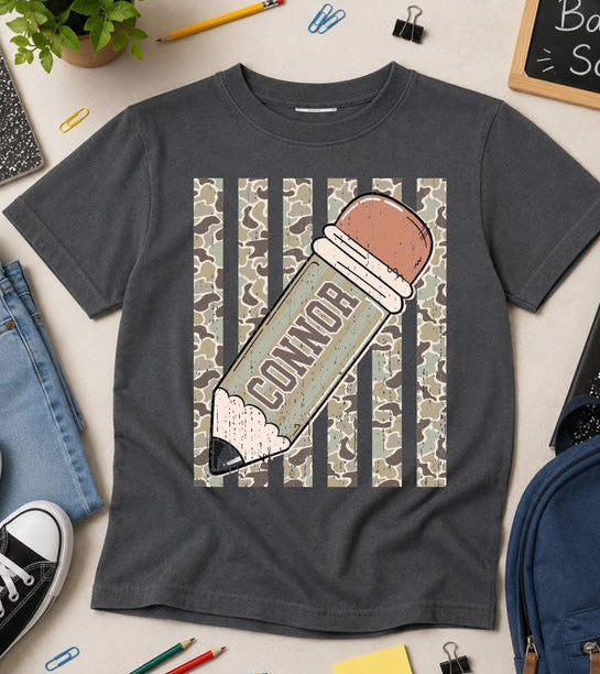 Gray t-shirt with a pencil graphic and 'CONNOR' text, surrounded by school supplies on a light surface.